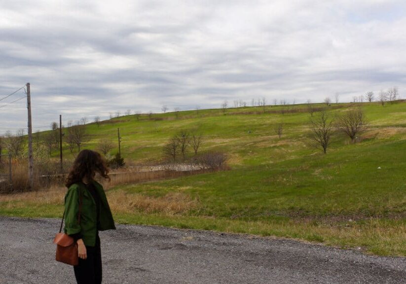 We see a woman in a green jacket looking at the grade of the mound at Freshkills Park.
